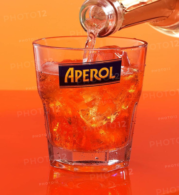 Aperol on ice cube cubes in an aperitif tumbler against an orange background is topped up with soda water