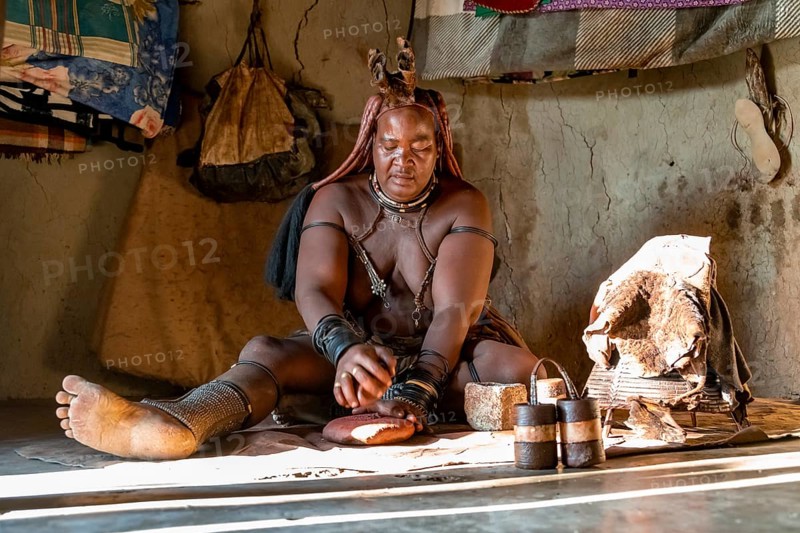 Traditional Himba woman making ochre body paint from fat and ochre, interior of the first woman's mud hut in a Himba village, traditional containers for ochre paint and woven frame with leather for body care with smoke, near Opuwo, Kaokoveld, Kunene, Namibia, Africa