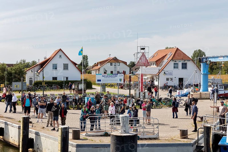 People gather at the harbour next to buildings and bicycles, a flag in the background, Rügen, Hiddensee