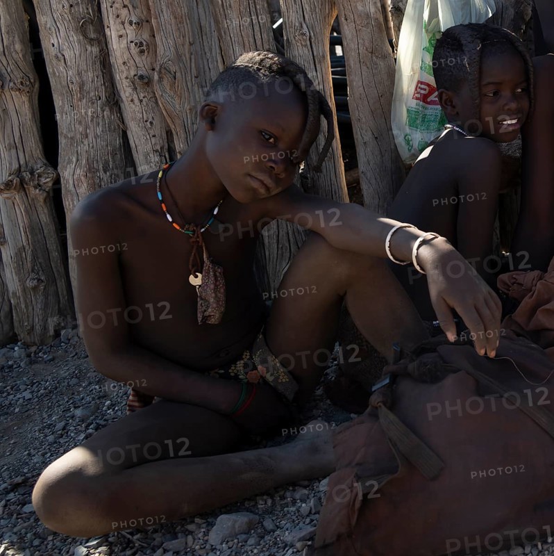 Himba girl with braided pigtails sitting in the shade near a hut, near Opuwo, Kaokoveld, Kunene, Namibia, Africa