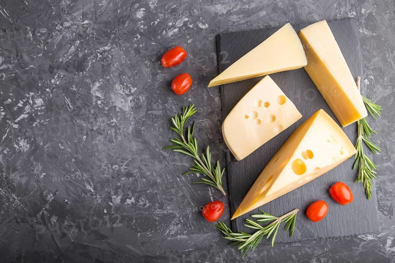 Various types of cheese with rosemary and tomatoes on black slate board on a black concrete background. Top view, flat lay, copy space