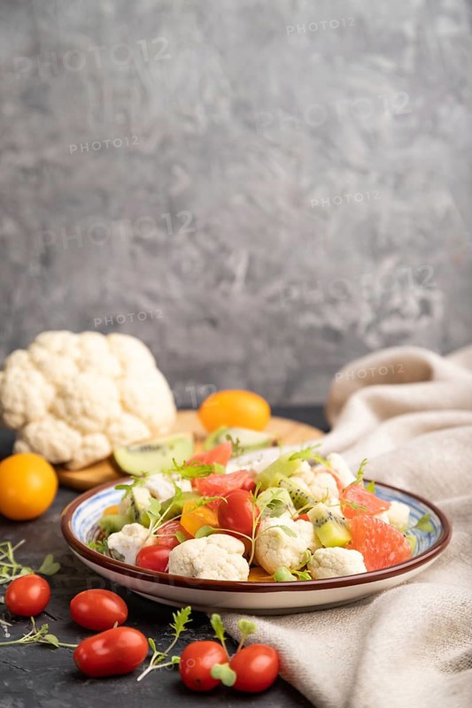 Vegetarian salad of cauliflower cabbage, kiwi, tomatoes, microgreen sprouts on black concrete background and linen textile. Side view, copy space