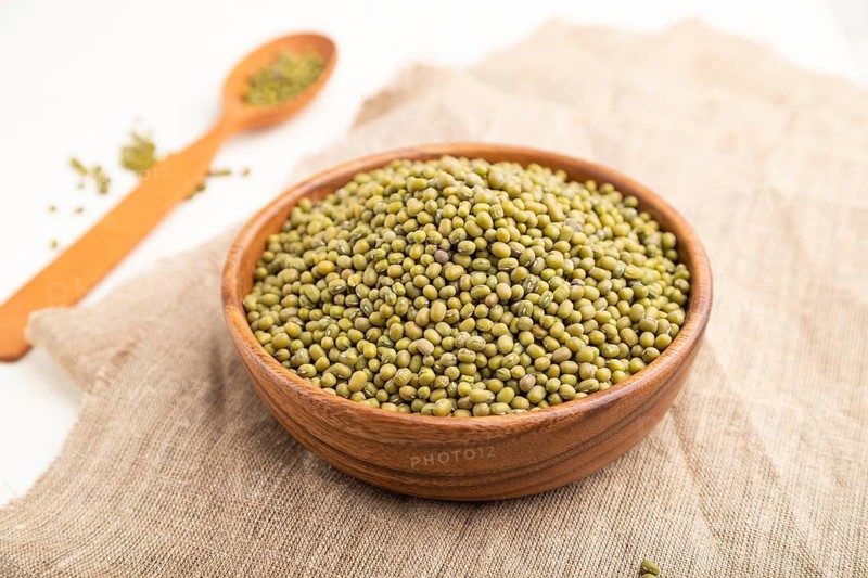 Wooden bowl with raw green mung bean and wooden spoon on a white background and linen textile. Side view, close up, selective focus