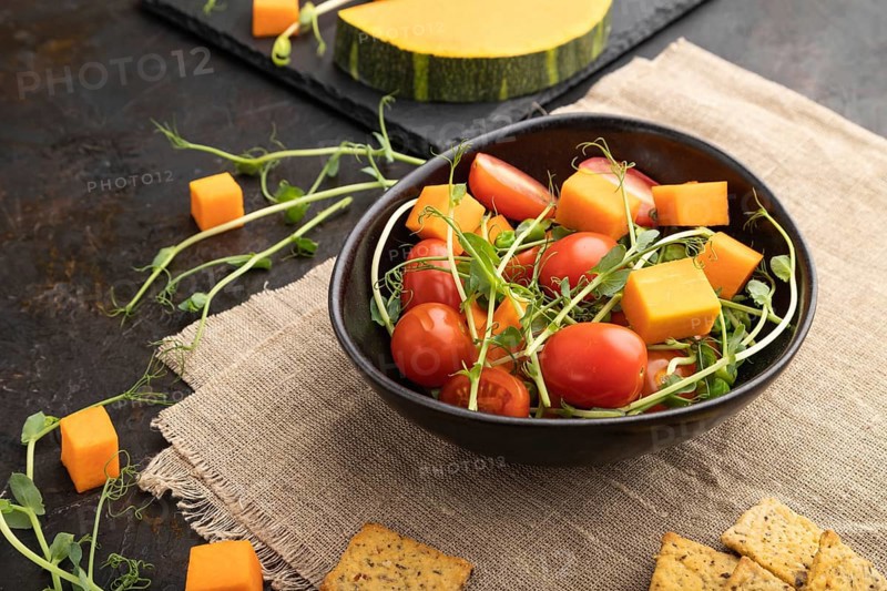 Vegetarian vegetable salad of tomatoes, pumpkin, microgreen pea sprouts on black concrete background and linen textile. Side view, close up