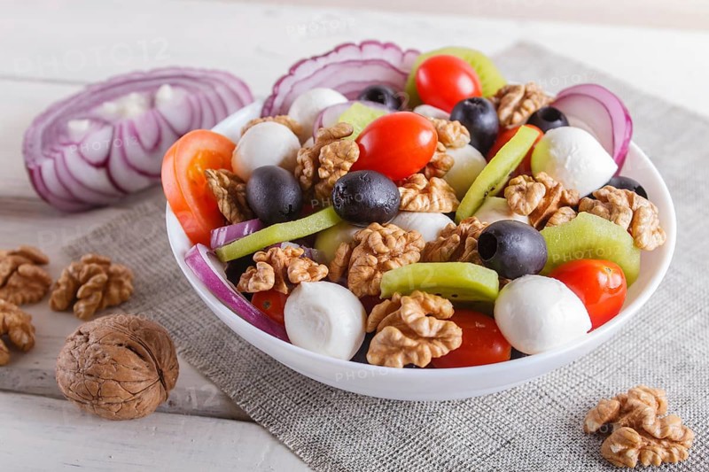 Salad with fresh cherry tomatoes, mozzarella cheese, olives, kiwi, onion and walnuts on white wooden background, copy space, close up