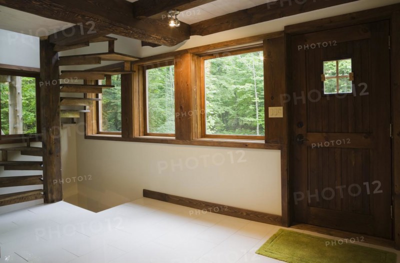Brown stained cherry wood spiraling staircase with thin flat wooden steps attached to pine wood post and front entrance door with square window inside Scandinavian hybrid style log cabin home, Quebec, Canada, North America