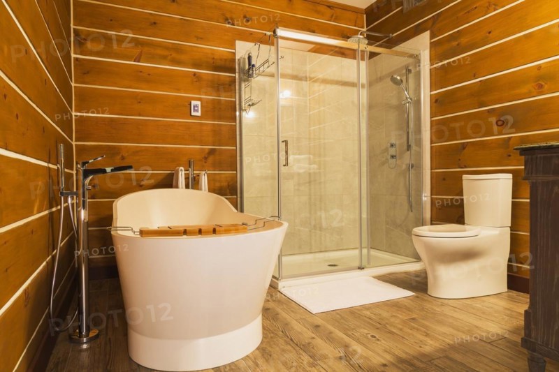 Freestanding bathtub with high tank toilet and clear glass shower stall in en suite with imitation wood ceramic tile flooring inside a milled Eastern white pine timber and flat log profile home, Quebec, Canada, North America