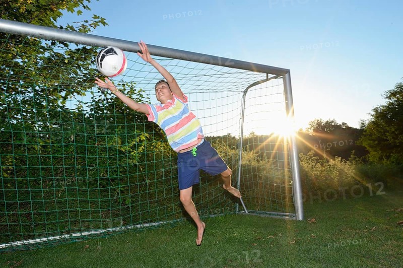 A boy jumping to save a soccer ball as a goalkeeper, with the sun shining in the background, Bavaria