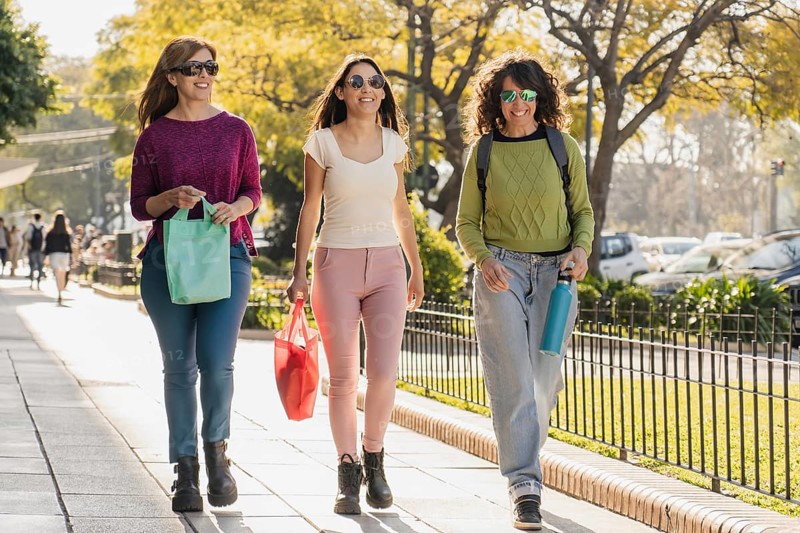 A group of three women strolling happily outdoors, carrying shopping bags on a sunny day, dressed in casual outfits and sunglasses, enjoying their time together