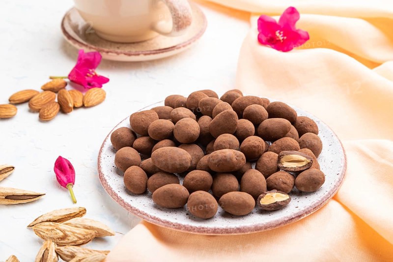 Almond in chocolate dragees on ceramic plate and a cup of coffee on white concrete background and orange linen textile. Side view, close up