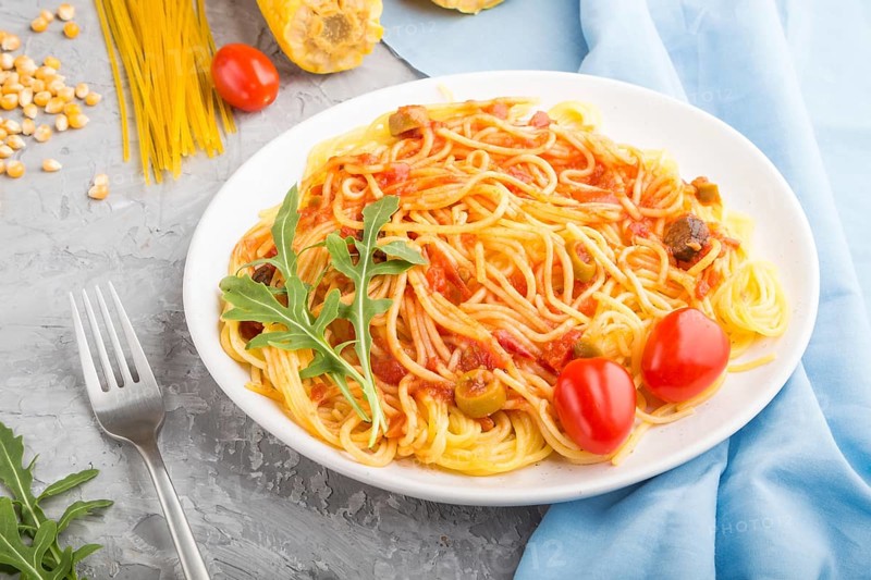 Corn noodles with tomato sauce and arugula on a gray concrete background and blue textile. Side view, close up