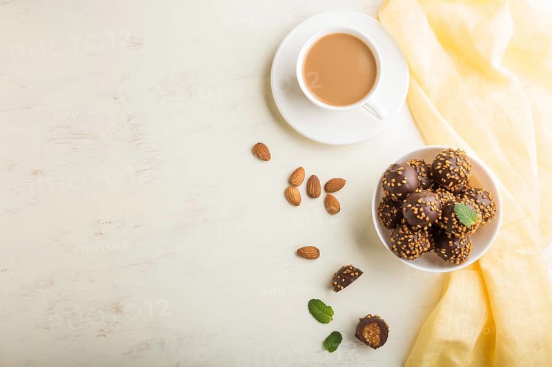 Chocolate caramel ball candies with almonds and a cup of coffee on a white wooden background and yellow textile. top view, flat lay, copy space