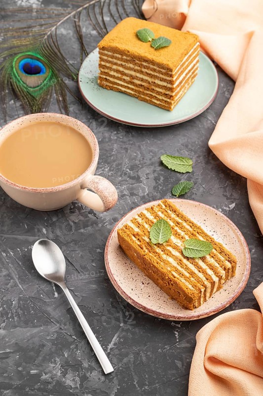 Homemade honey cake with milk cream and mint with cup of coffee on a black concrete background and orange textile. Side view, close up