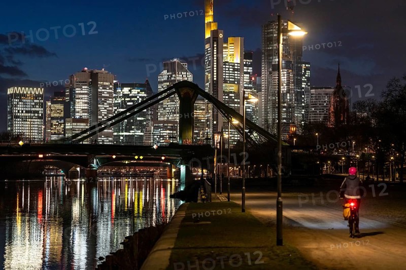 Skyline of the city centre of Frankfurt am Main, cyclist with light on the cycle path, pavement, promenade along the river Main, dusk, Hesse, Germany raft bridge in the background