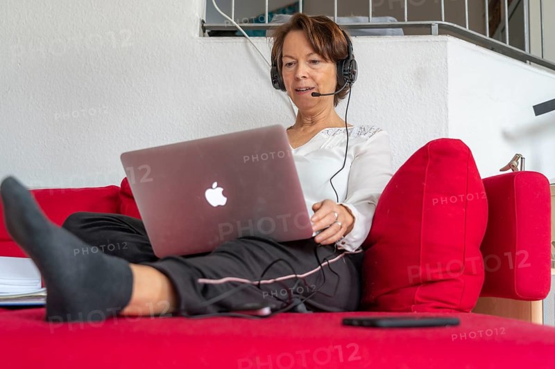 Woman, mid-50s, works from home, with laptop and communicates with colleagues via headset, home office, on the sofa
