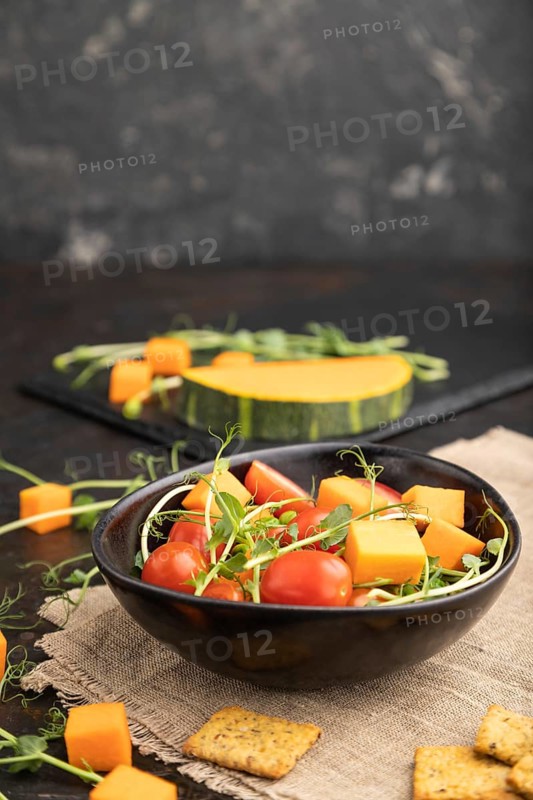 Vegetarian vegetable salad of tomatoes, pumpkin, microgreen pea sprouts on black concrete background and linen textile. Side view, copy space