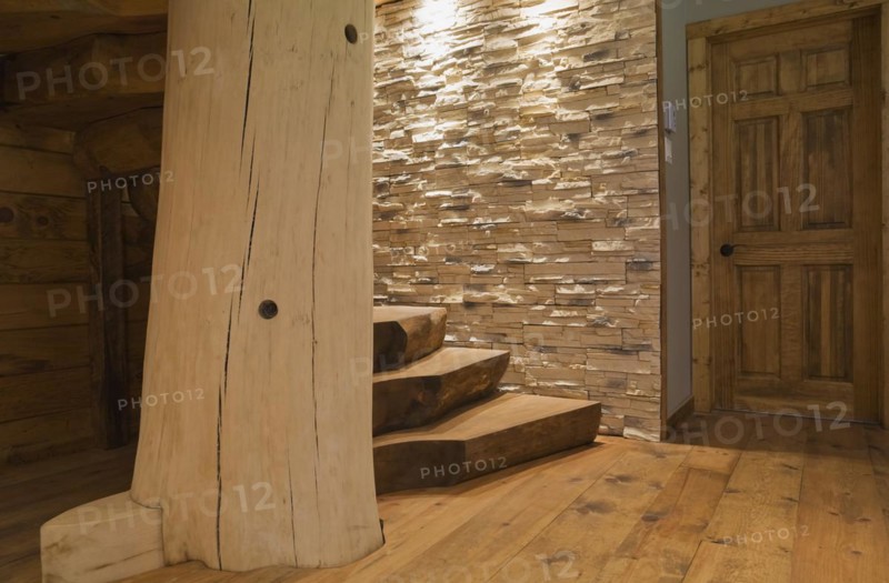 Door and twisting wooden staircase with brown stained half log steps attached to large bleached supporting central tree trunk post and illuminated lights shining on tan natural stone wall in basement leading to ground floor inside handcrafted Eastern white pine Scandinavian log cabin home, Quebec, Canada, North America