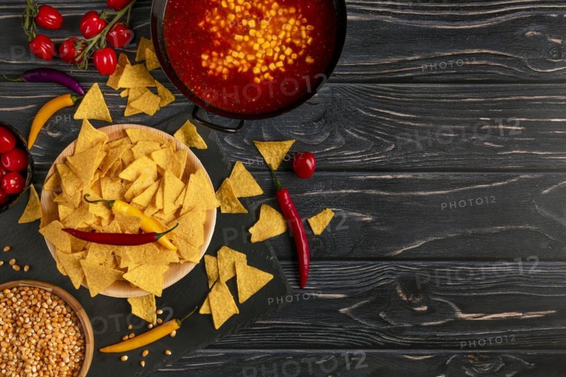 Tortilla chips next to a pan with chilli con carne, peppers and tomatoes on a dark wooden background