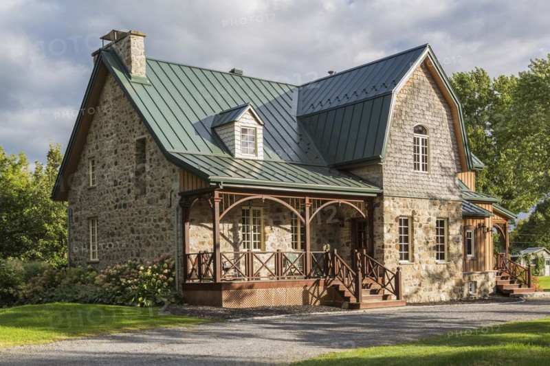 Old 1826 Canadiana cottage style fieldstone house facade with vertical wood plank cladding, cedar shingles and green standing seam sheet metal roof in late summer, Quebec, Canada, North America
