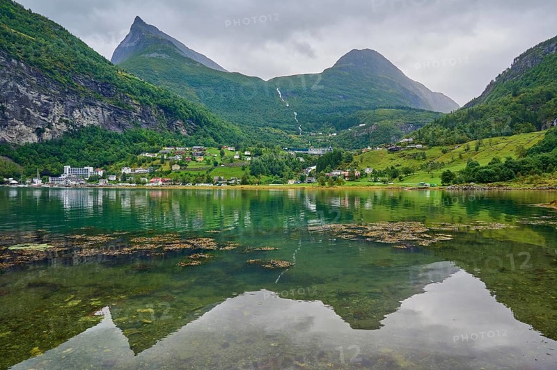 Mountainous landscape with a calm fjord reflecting the surrounding mountains and village, surrounded by green nature and under a cloudy sky, Geiranger, Geiranger Fjord, Stranda, Romsdal, Norway, Europe