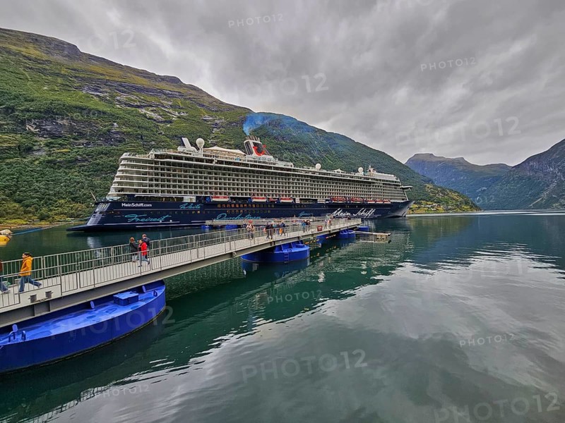 A large cruise ship, Mein Schiff 6, lies in the fjord, surrounded by green mountains under a cloudy sky, with catwalk and tourists, Geiranger, Geiranger Fjord, Stranda, Romsdal, Norway, Europe