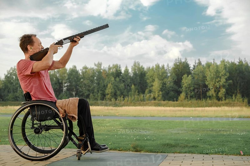 Senior man with paralysed legs in wheelchair aiming double-barreled shotgun at an outdoor shooting range