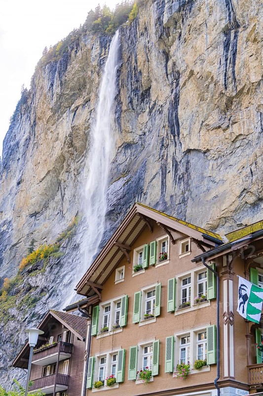 An impressive waterfall cascades down next to a wooden house with green shutters. A steep rock face dominates the scene, Lauterbrunnen, Switzerland, Europe