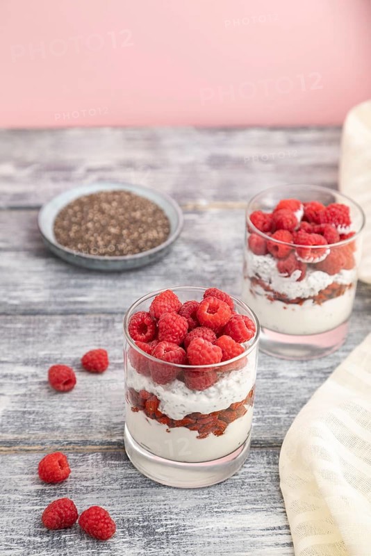Yogurt with raspberry, goji berries and chia seeds in glass on gray wooden background and linen textile. Side view, close up, selective focus