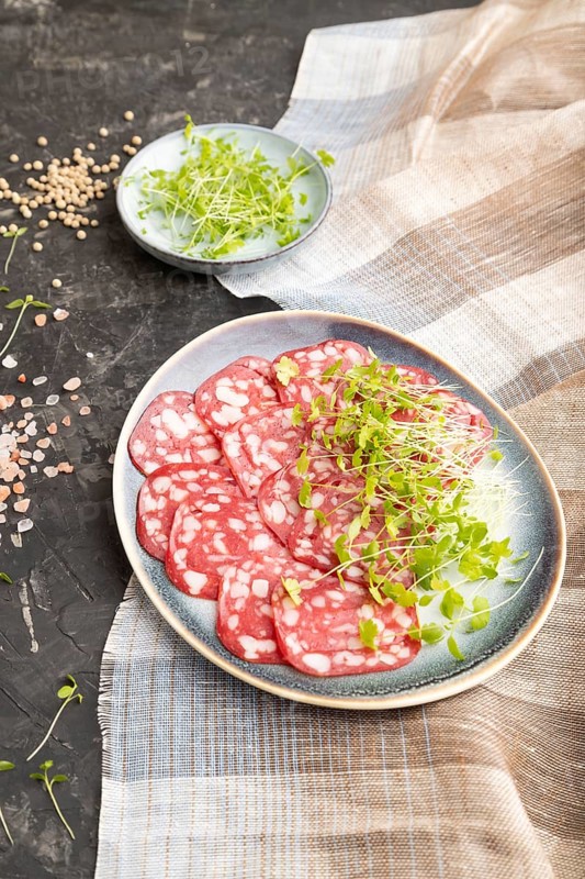 Slices of smoked cervelat salami sausage with spinach microgreen, salt and pepper on black concrete background and beige textile. Side view, close up