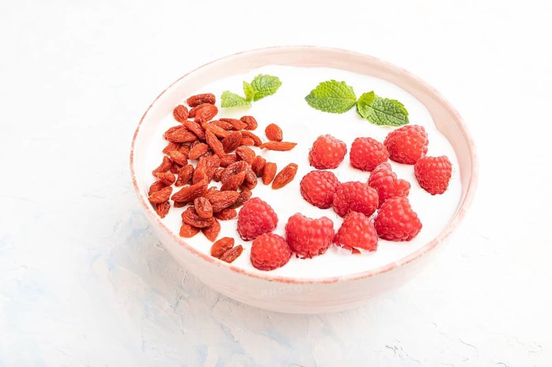 Yogurt with raspberry and goji berries in ceramic bowl on white concrete background. Side view, close up