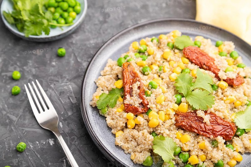 Quinoa porridge with green pea, corn and dried tomatoes on ceramic plate on a gray concrete background and yellow textile. Side view, close up, selective focus