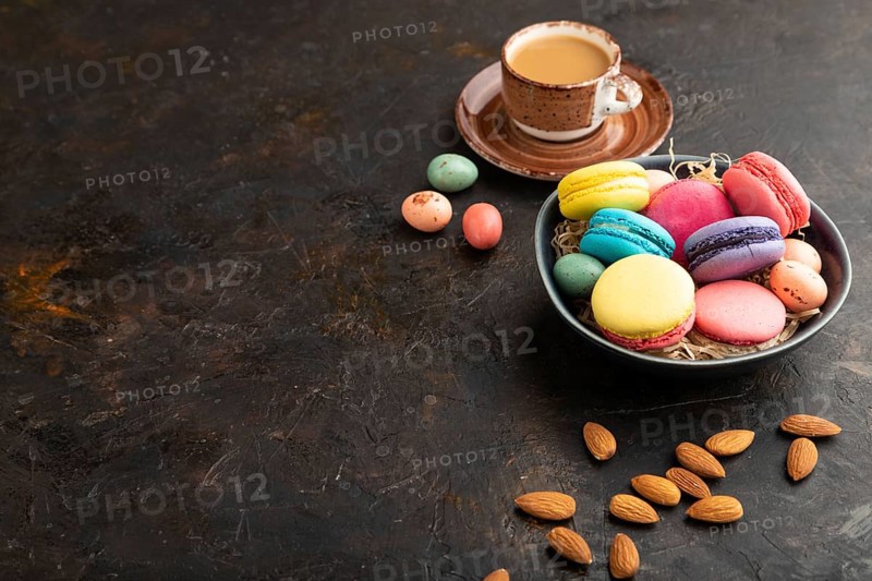 Multicolored macaroons and chocolate eggs in ceramic bowl, cup of coffee on black concrete background. side view, copy space, still life. Breakfast, morning, concept