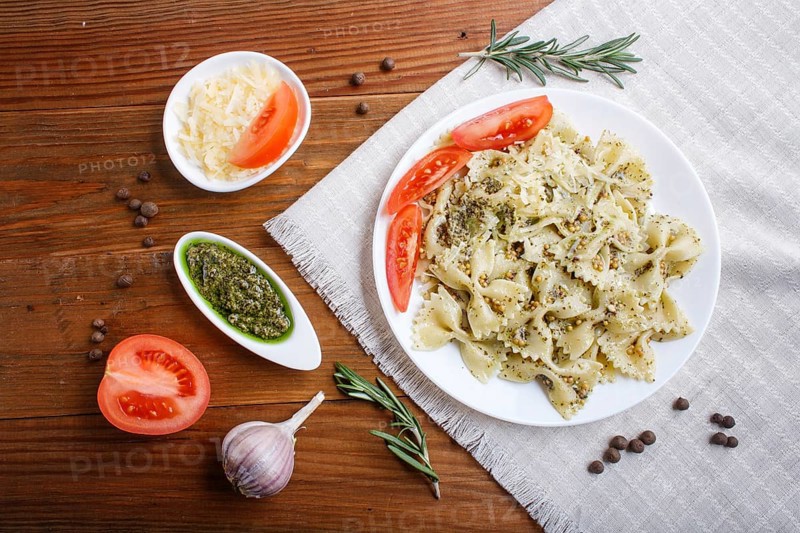 Farfalle pasta with pesto sauce, tomatoes and cheese on a linen tablecloth on brown wooden background. top view, close up