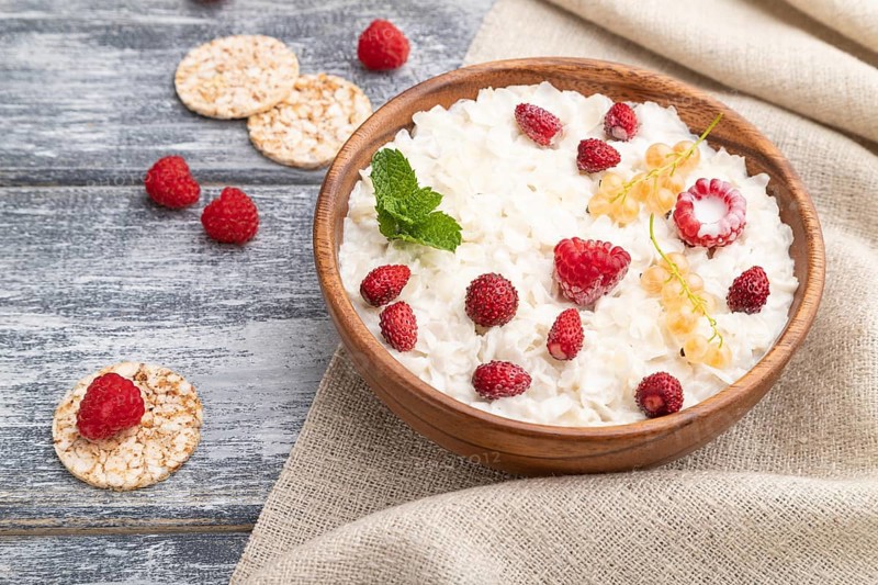Rice flakes porridge with milk and strawberry in wooden bowl on gray wooden background and linen textile. Side view, close up