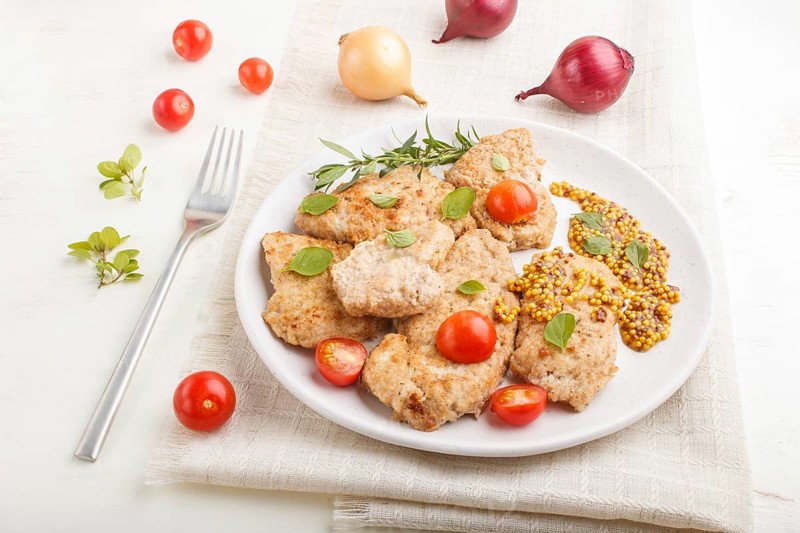 Fried pork chops with tomatoes and herbs on a white ceramic plate on a white wooden background and linen textile. side view, close up