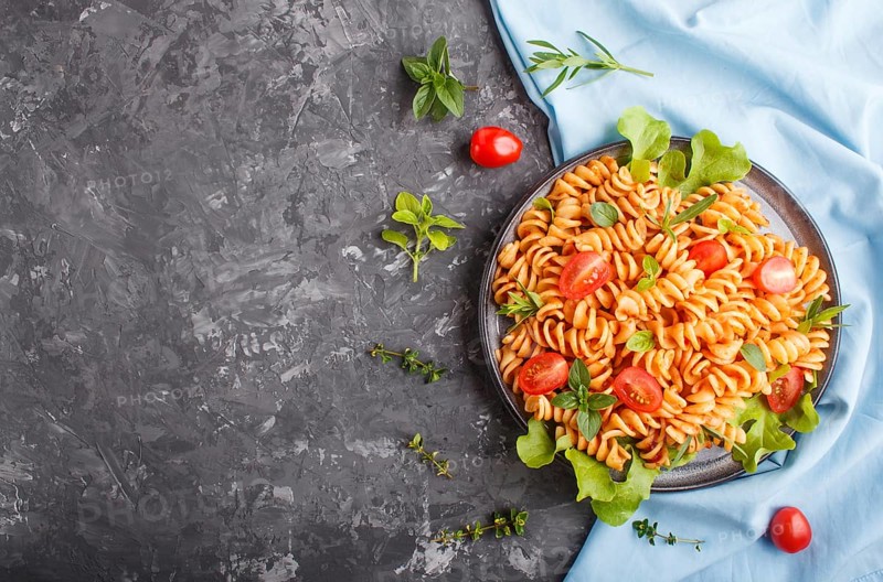 Fusilli pasta with tomato sauce, cherry tomatoes, lettuce and herbs on a black concrete background with blue textile. top view, flat lay, copy space