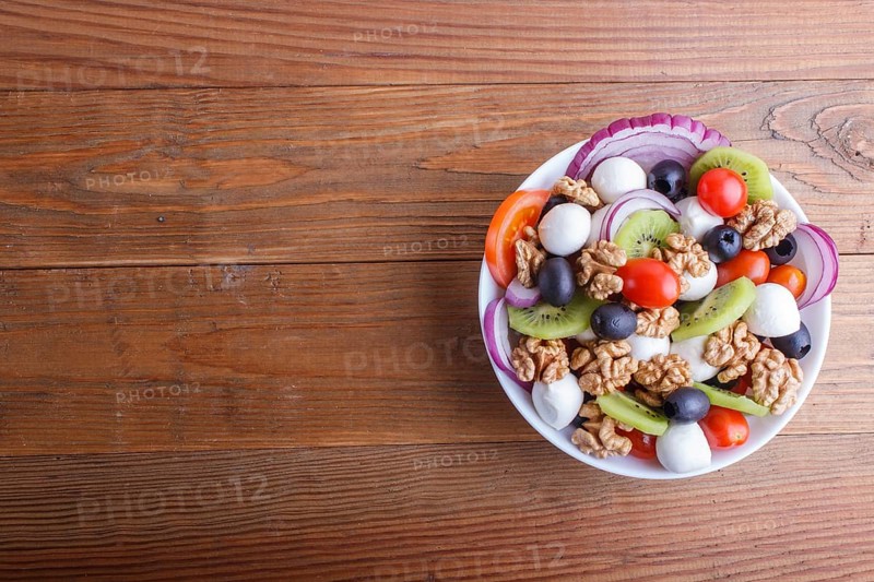 Salad with fresh cherry tomatoes, mozzarella cheese, olives, kiwi, onion and walnuts on brown wooden background, copy space, top view