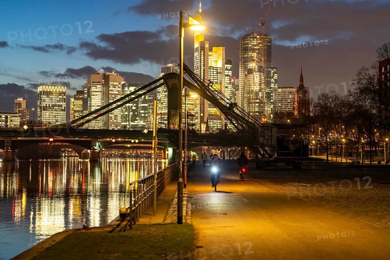 Skyline of the city centre of Frankfurt am Main, cyclist with light on the cycle path, pavement, promenade along the river Main, dusk, Hesse, Germany raft bridge in the background