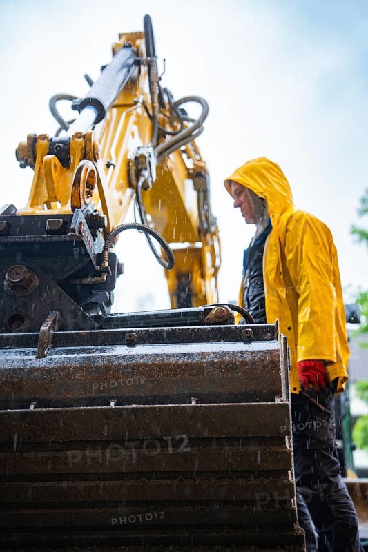 Construction worker in yellow rain jacket working with large construction machine in the rain, construction site Hermann Hesse railway, Calw, Black Forest, Germany, Europe
