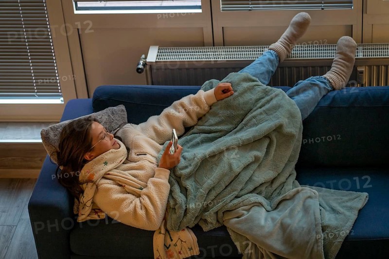 Symbolic image energy saving, cold winter, energy crisis, cold flat, mother and daughter lying on the sofa, warmly dressed, candlelight, heating on to a minimum, warming their feet on the radiator