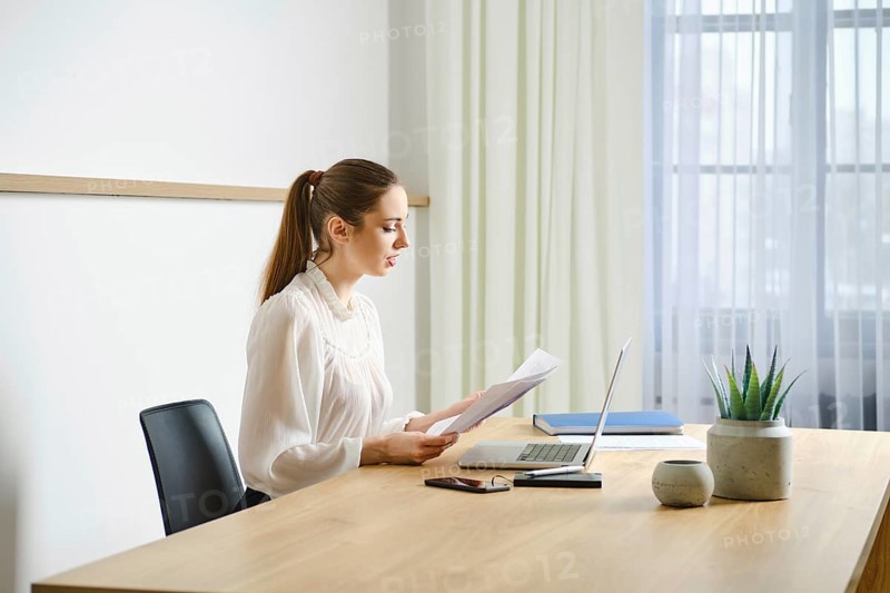 A focused professional woman realtor is reviewing a paper document at her clean, organized workspace in a bright, modern office