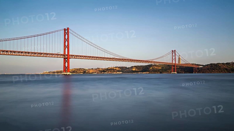 Serene dusk view of a red suspension bridge over calm blue water with reflections, Lissabon, 25 de Abril Bridge