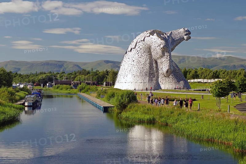 Monumental statue of two horses beside a canal with boats and people, surrounded by greenery and woodland under a blue sky, sculptures, Kelpies, Falkirk, Scotland, United Kingdom, Europe