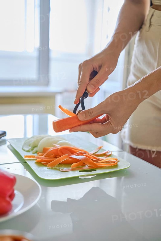 An unrecognizable woman peeling carrots on a green cutting board in a sunlit kitchen. Slices of carrot and onion are neatly arranged, indicating meal preparation in a vibrant, homey space