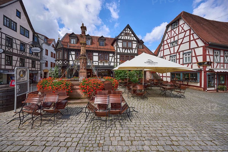 Market fountain with geraniums, half-timbered houses and outdoor areas of a restaurant with parasols, tables and chairs on the market square in Michelstadt, Odenwald, Odenwaldkreis, Hesse, Germany, Europe