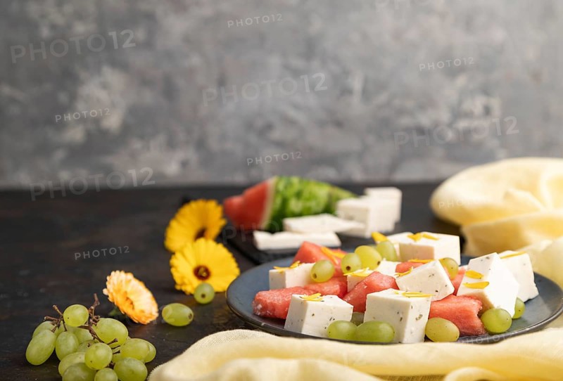 Vegetarian salad with watermelon, feta cheese, and grapes on blue ceramic plate on black concrete background and yellow linen textile. Side view, copy space, selective focus
