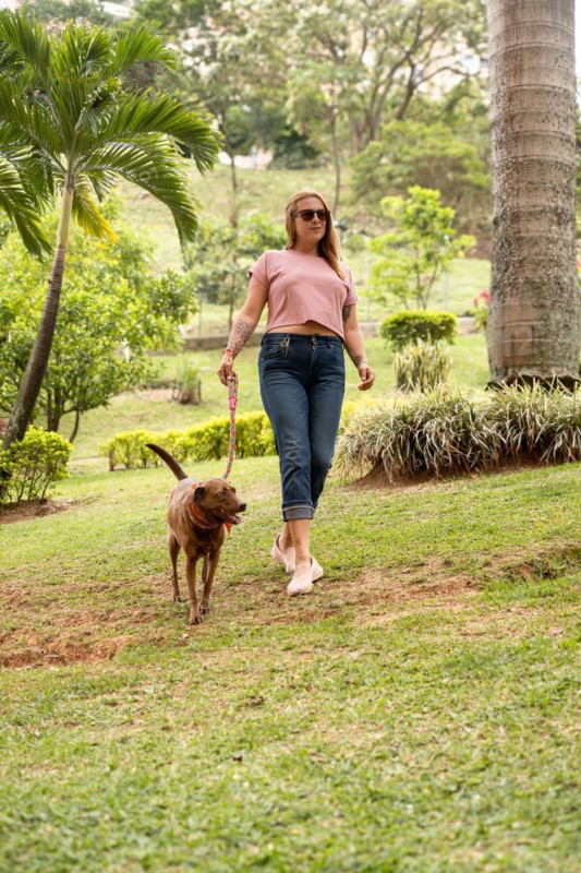 Vertical shot of a happy woman walking on the grass with her pet beside her outdoors. The woman is wearing jeans, sneakers, and sunglasses, and holding her dog on a leash