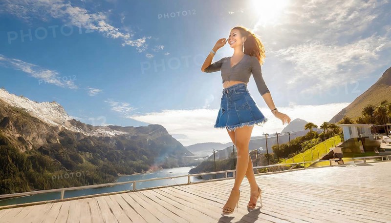 Woman in a denim skirt and sandals stands on a sunny wooden terrace with a view of mountains and a lake, KI generated, AI generated