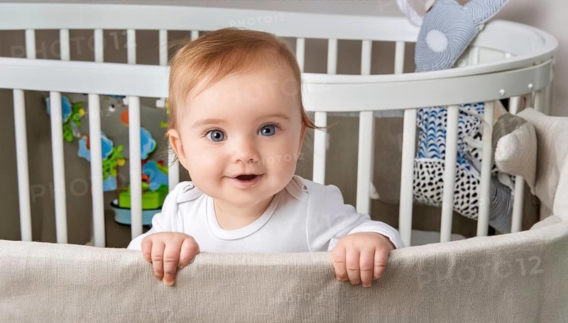 A happy baby stands smiling in a cot with toys in the background, AI generated, AI generated