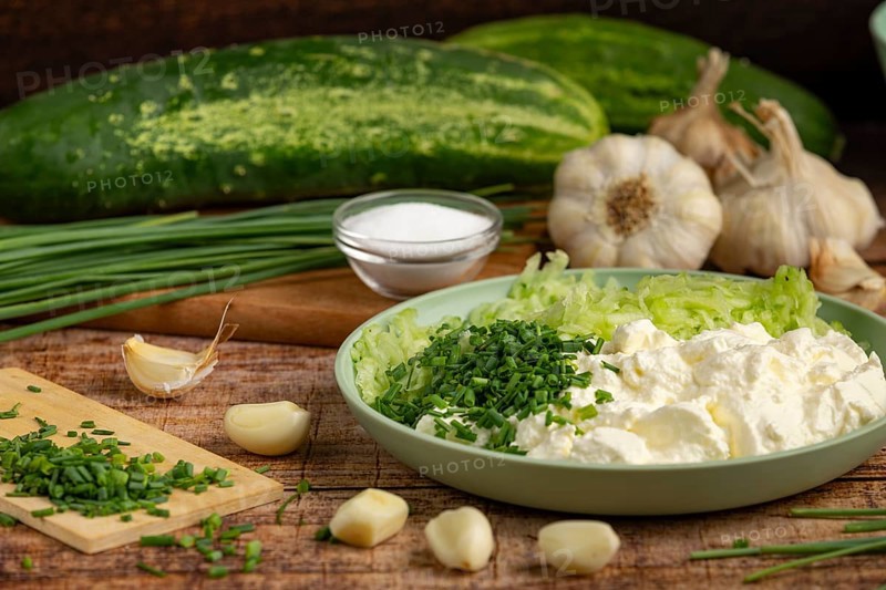 Fresh ingredients such as whole and grated cucumbers, garlic, chopped chives and sour cream on a wooden table